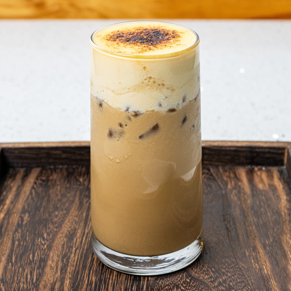 Layered iced coffee topped with creamy foam and toasted spice, served in a clear glass on a wooden tray.