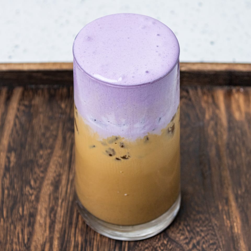 Layered golden smoothie topped with thick purple foam, served in a clear glass on a wooden table.