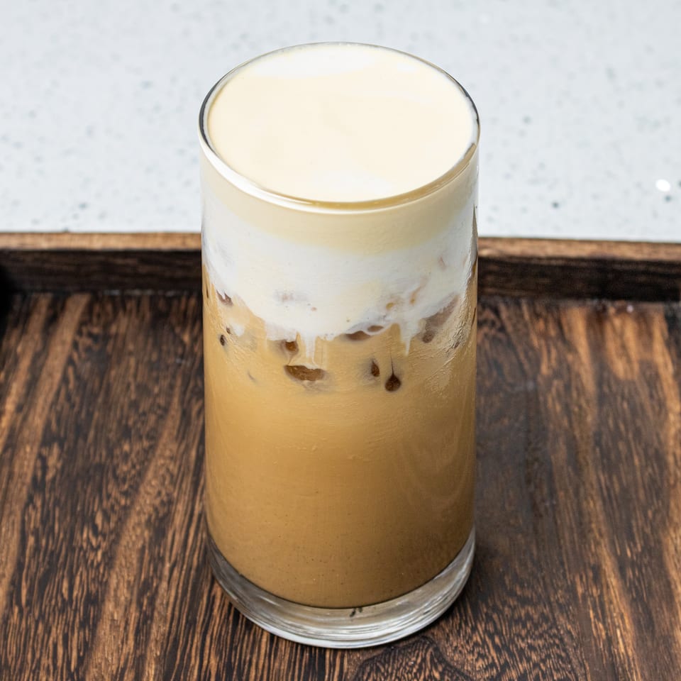 Layered iced coffee topped with creamy foam, served in a clear glass on a dark wooden tray.
