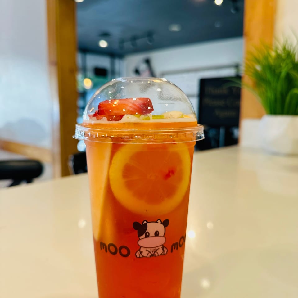 Orange fruit drink with strawberry and citrus slices under a clear dome lid, served in a cow-branded cup at a café.