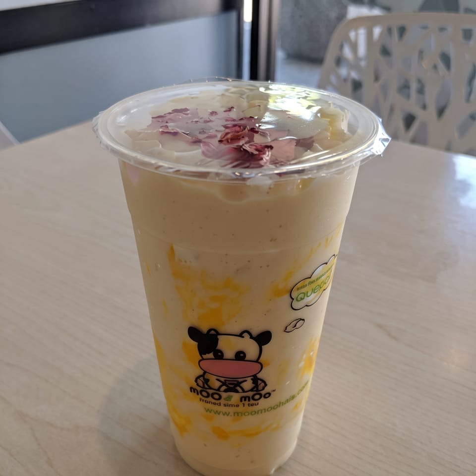 Moo Moo-branded cup of creamy mango milk tea topped with dried rose petals, sitting on a wooden restaurant table.