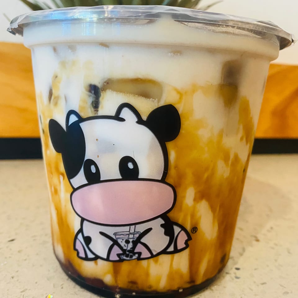Sealed plastic cup of a creamy caramel-colored beverage featuring a cartoon cow graphic, resting on a café counter.