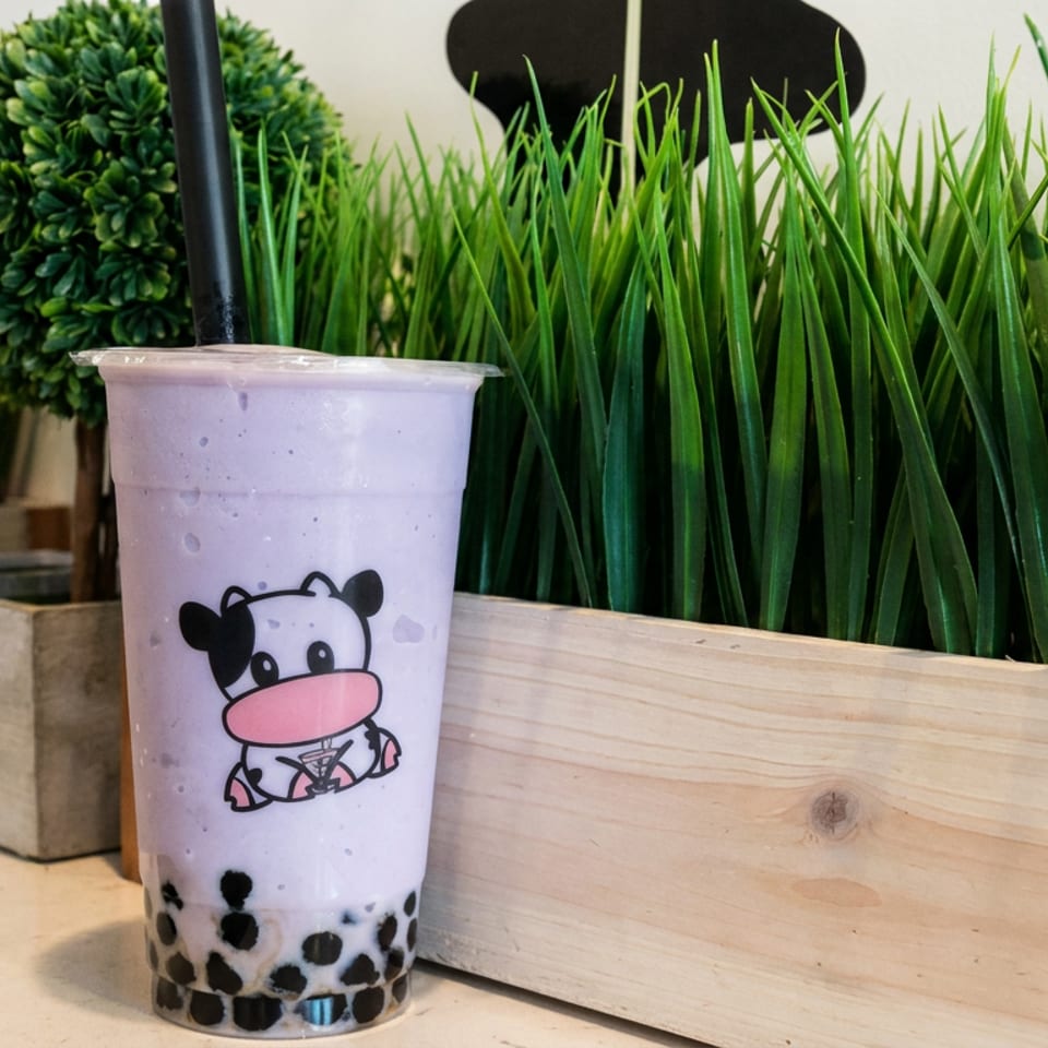 Lavender milk tea with black tapioca pearls in a cartoon cow cup, displayed before a planter of tall green grass.