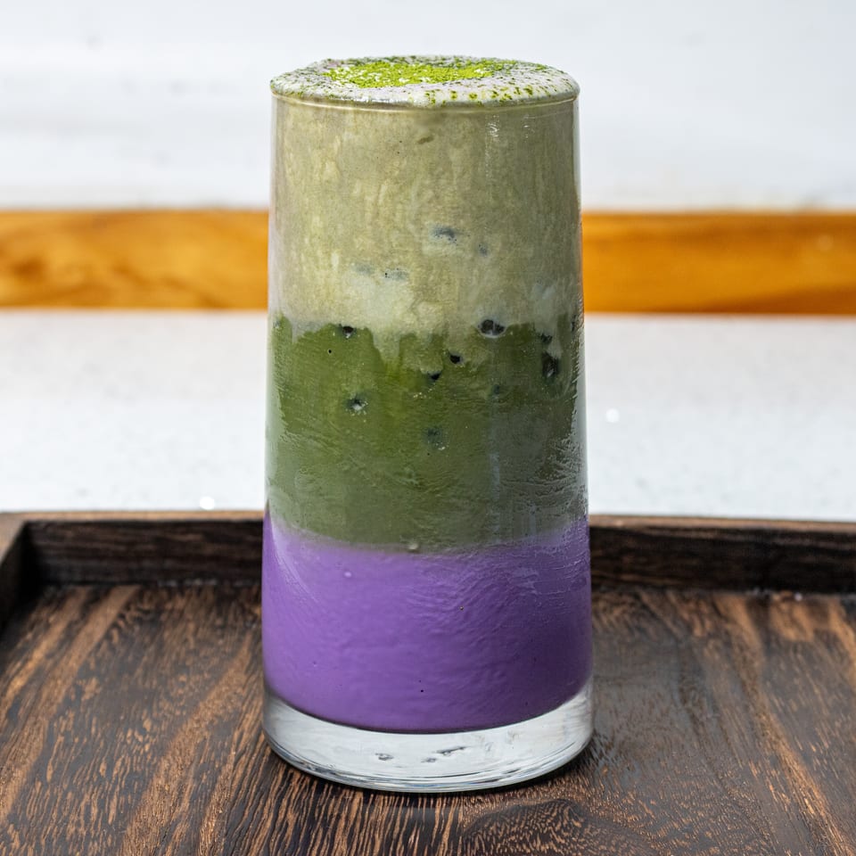 Layered purple and green matcha drink topped with green powder, served in a clear glass on a wooden tray.