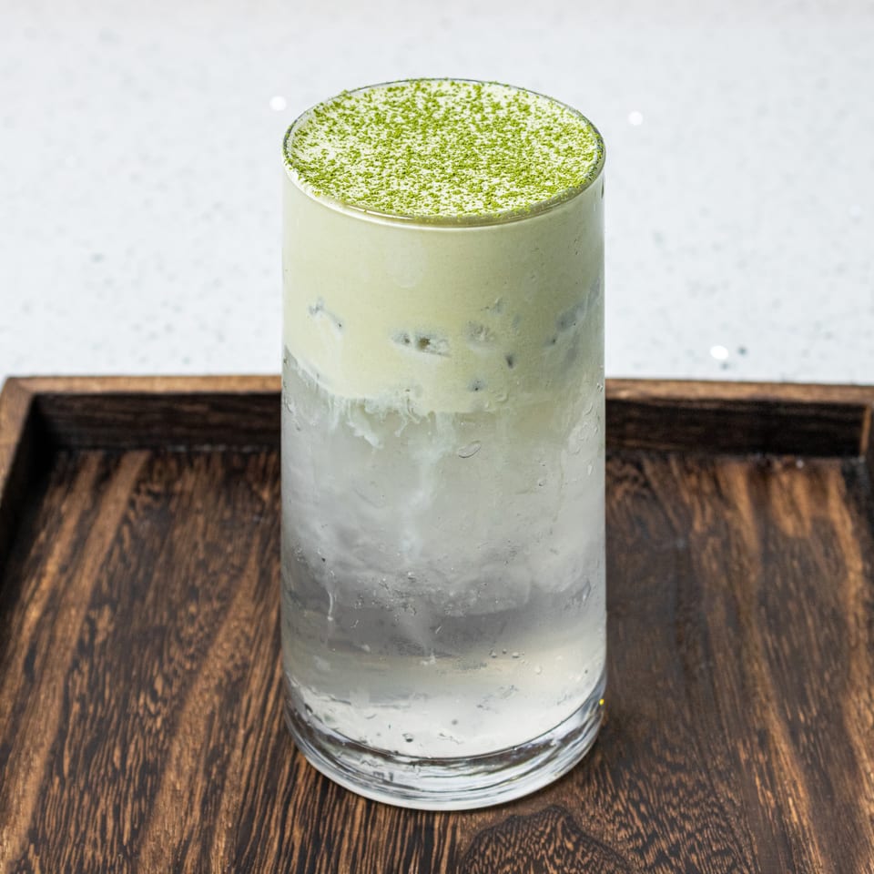 Iced matcha latte topped with green matcha powder in a clear glass on a dark wooden tray.
