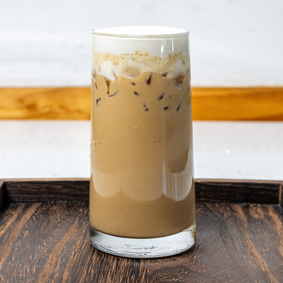 Tall glass of iced coffee topped with creamy foam, served on a dark wooden tray.